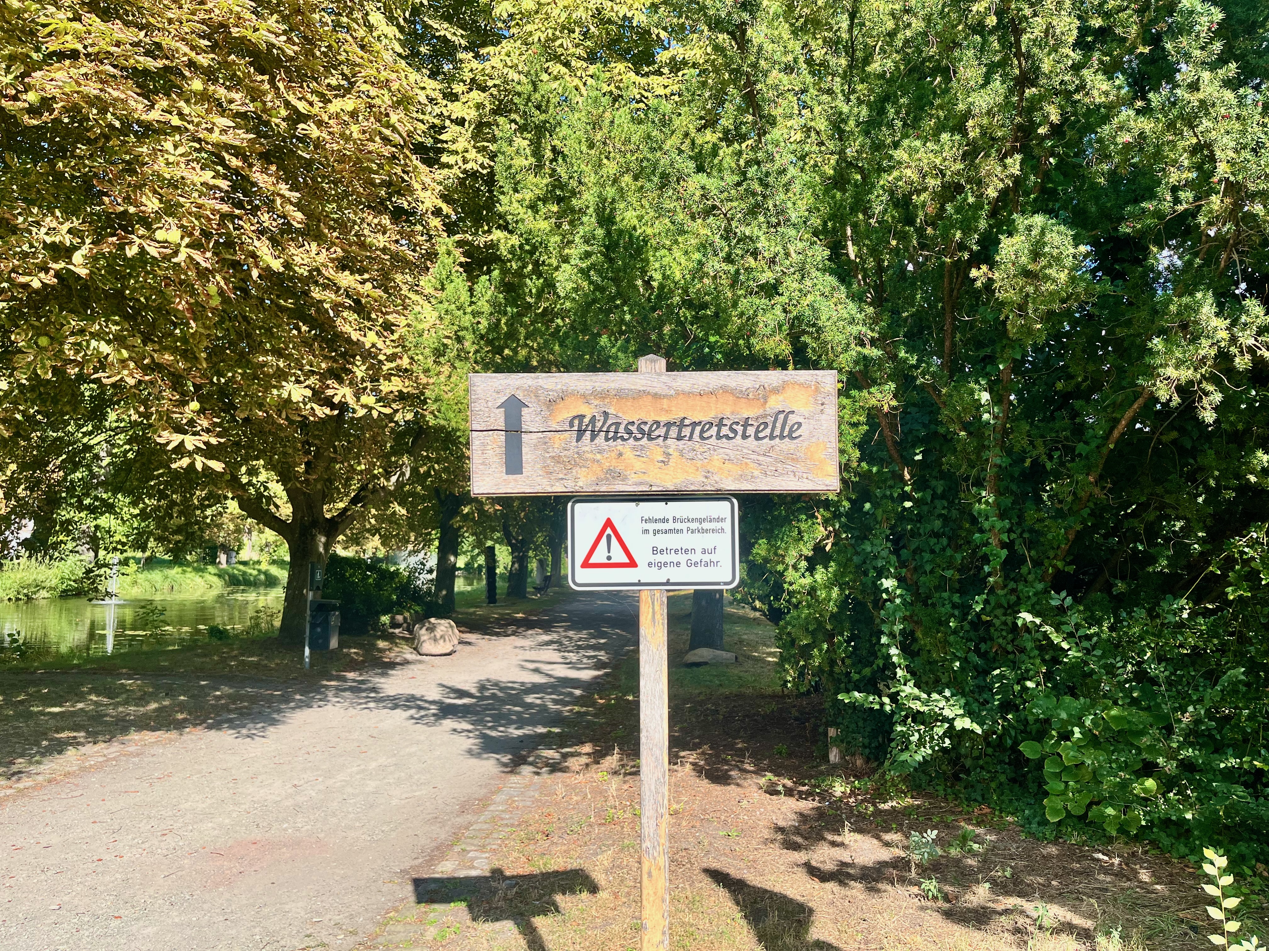 Holzschild mit Pfeil zur Wassertretstelle an einem schattigen Weg im grünen Kurpark. Darunter warnt ein Schild vor fehlenden Brückengeländern und dem Betreten auf eigene Gefahr. Links ist ein Gewässer zu sehen.