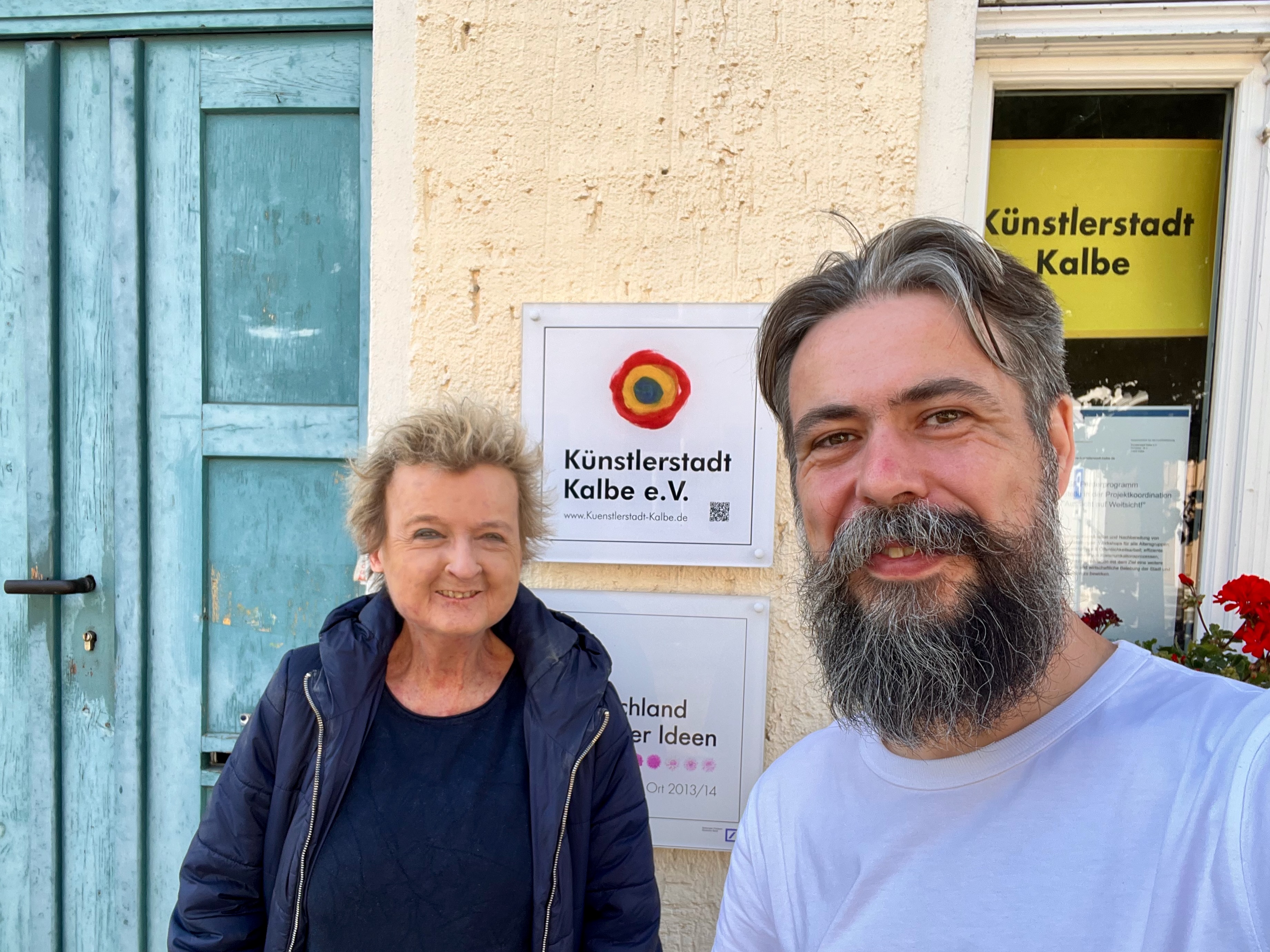 Corinna Köbele und Tobias Kremkau lächeln vor dem Eingang der Künstlerstadt Kalbe e. V. in die Kamera.