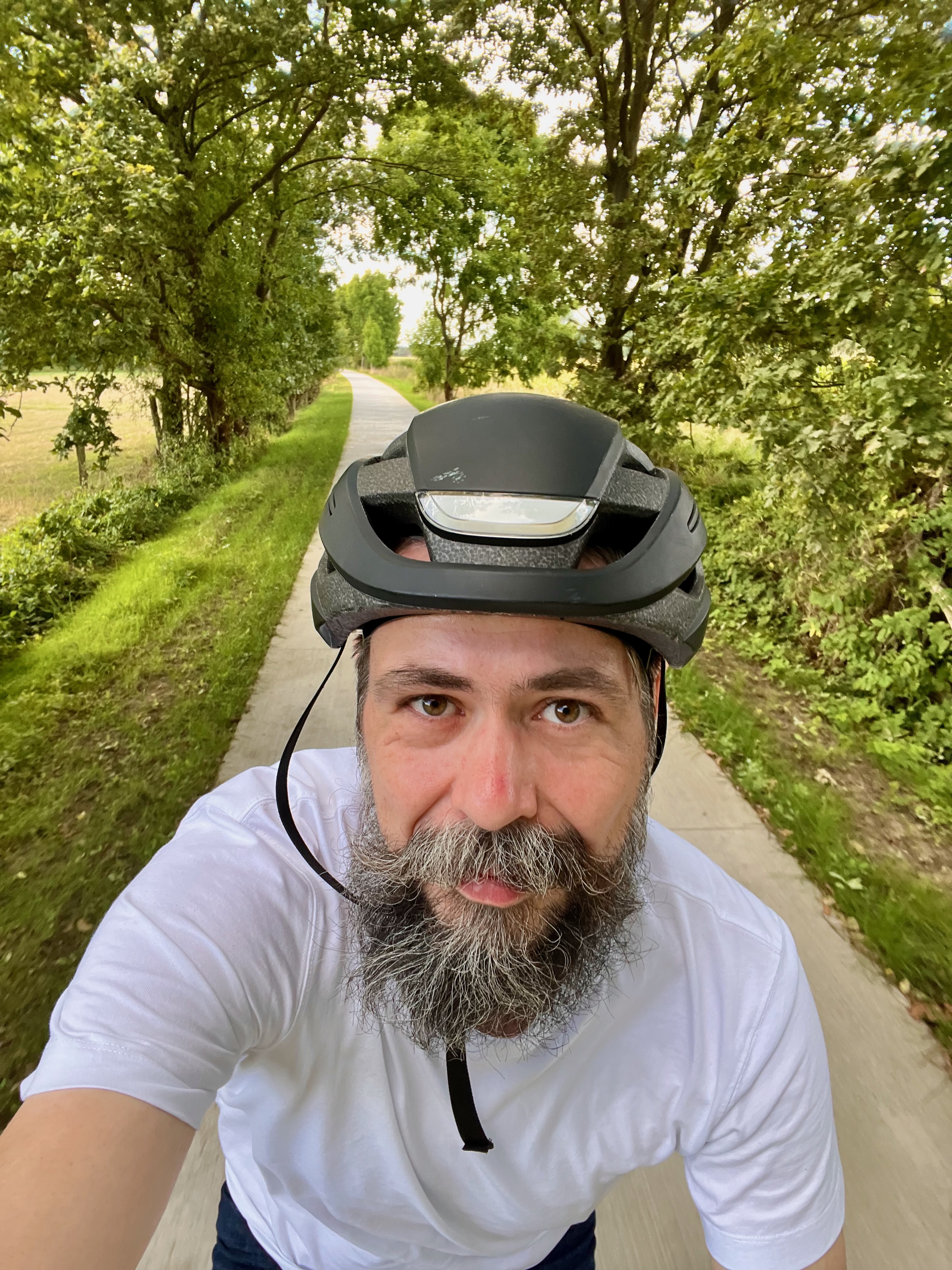 Selfie eines bärtigen Radfahrers mit schwarzem Fahrradhelm und weißem T-Shirt. Hinter ihm verläuft ein schmaler, heller Radweg durch eine dicht begrünte Baumallee.