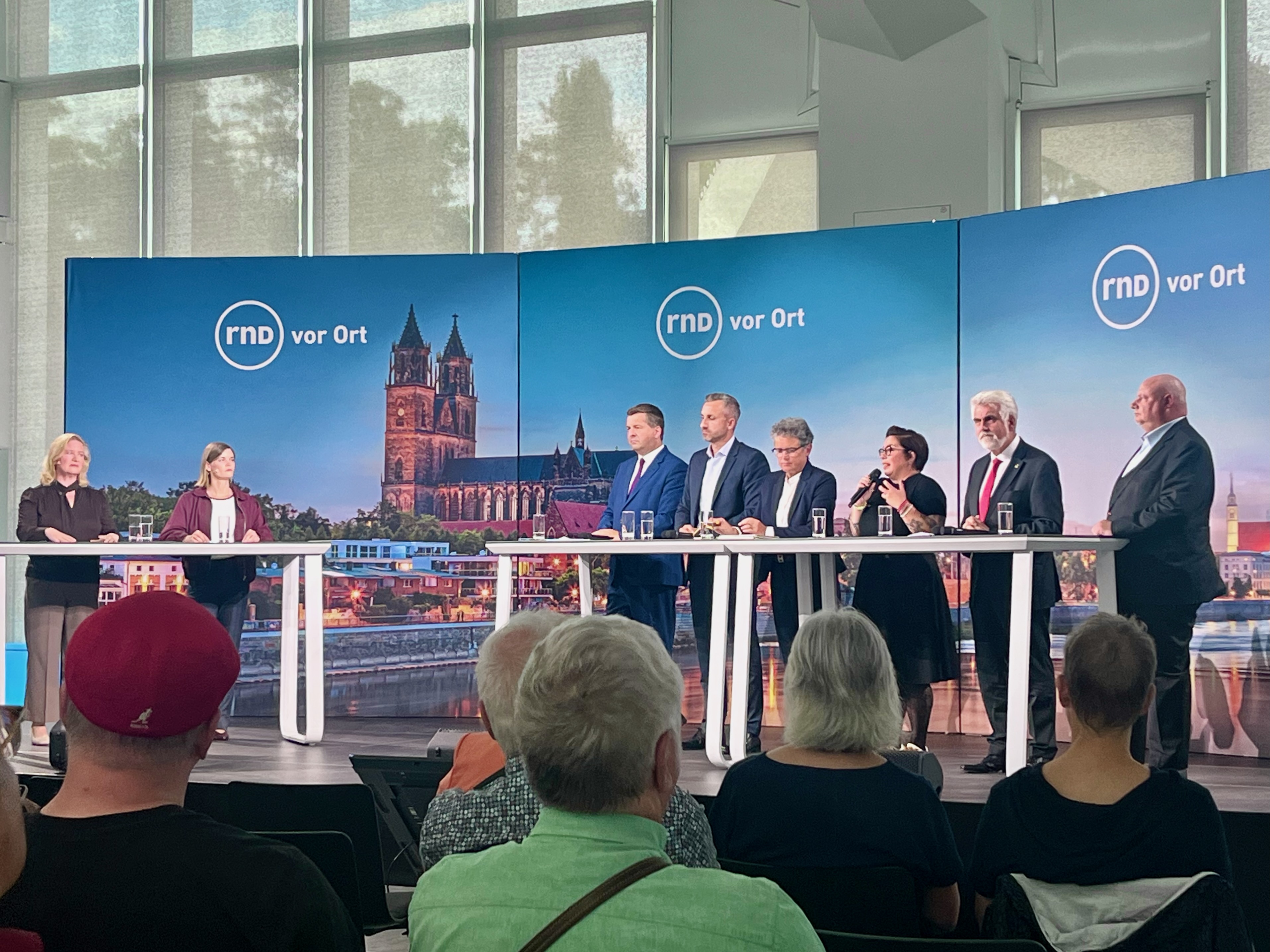 Zwei Moderatorinnen und sechs Politikerinnen und Politiker stehen bei einer Podiumsdiskussion des Redaktionsnetzwerks Deutschland auf einer Bühne hinter mehreren Stehtischen. Eine Frau spricht ins Mikrofon und gestikuliert. Im Vordergrund verfolgt das Publikum die Debatte; die Bühnenkulisse zeigt das Magdeburger Stadtpanorama mit dem Dom und die Aufschrift „rnd vor Ort“.